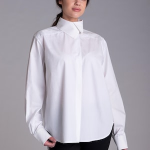 May include: A crisp white long-sleeved button-down shirt featuring a structured high collar, a concealed front placket, and wide cuffs. The shirt is made of a smooth, opaque fabric and is paired with simple black trousers, creating a clean look.
