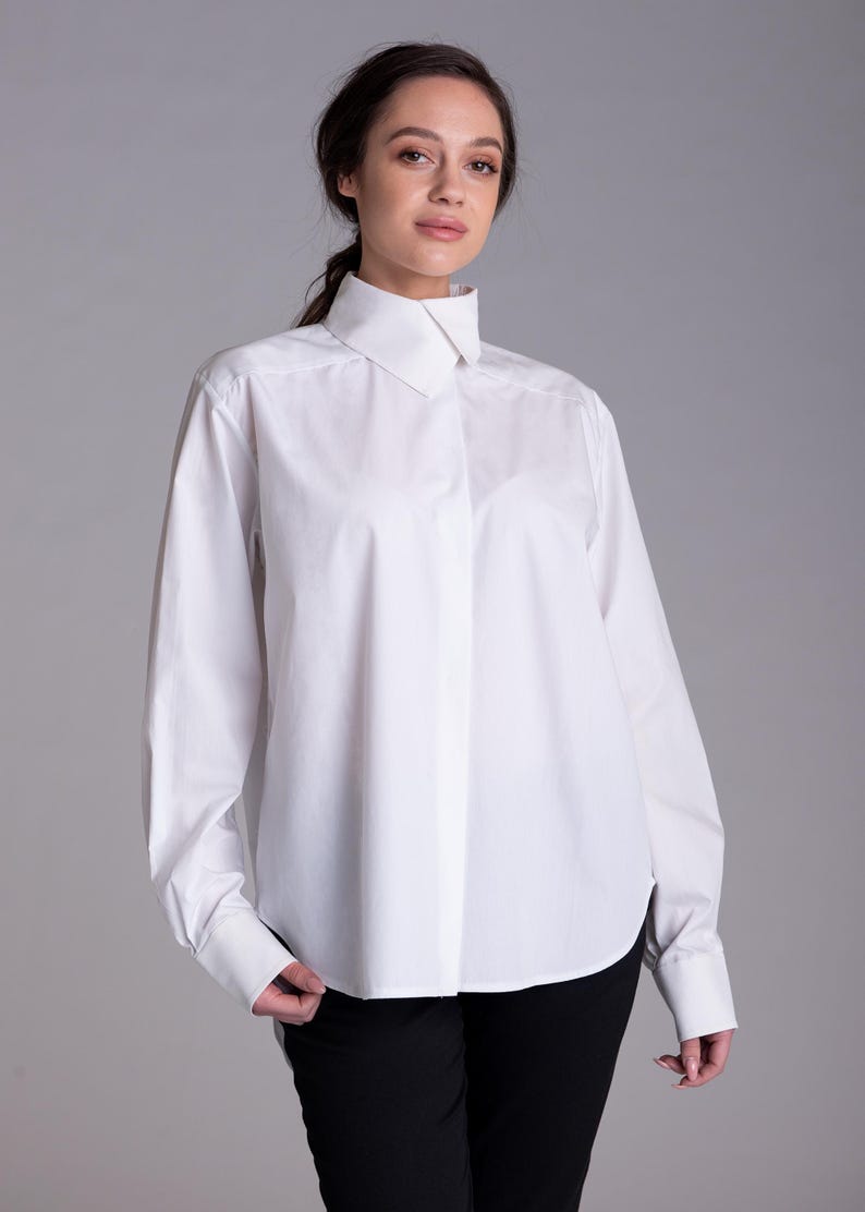 May include: A crisp white long-sleeved button-down shirt featuring a structured high collar, a concealed front placket, and wide cuffs. The shirt is made of a smooth, opaque fabric and is paired with simple black trousers, creating a clean look.