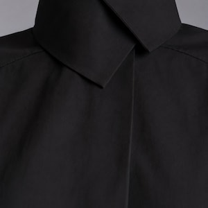 May include: A solid black shirt featuring a high, structured collar with a sharp, pointed fold that overlaps at the front. The garment is crafted from a smooth, matte fabric and displays clean, minimalist lines around the neck and shoulder area.