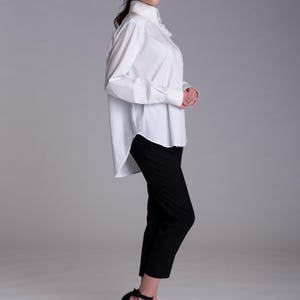 May include: A crisp white long-sleeved button-down shirt with a structured collar and cuffs, paired with slim-fit black trousers that end above the ankle. The shirt features a high-low hemline and a subtle ruffled detail along the front placket.