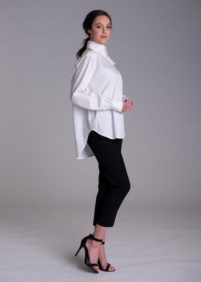 May include: A crisp white long-sleeved button-down shirt with a structured collar and cuffs, paired with slim-fit black trousers that end above the ankle. The shirt features a high-low hemline and a subtle ruffled detail along the front placket.