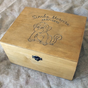 Op de afbeelding: Een houten doos met een scharnierend deksel en een metalen sluiting. De doos is gegraveerd met de naam "Simba Moreira" en een cartoon-aap.