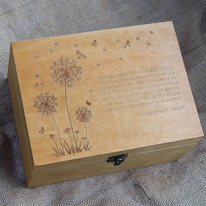 Op de afbeelding: Houten doos met een scharnierend deksel en een metalen sluiting. De doos is versierd met een gegraveerd ontwerp van paardenbloemen en vlinders. De quote "She was elusive. She was today. She was tomorrow. She was the faintest scent of a cactus flower, the flitting shadow of an elf owl. We did not know what to make of her. In our minds we tried to pin her to a cork board like a butterfly, but the pin merely went through and away she flew" - Jerry Spinelli, Stargirl is gegraveerd op de doos.