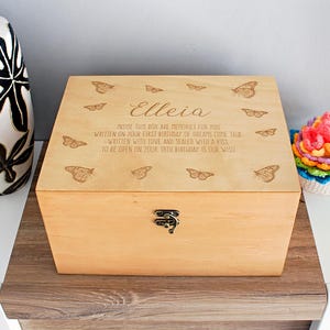 Puede incluir: Una caja de recuerdos de madera de color claro con tapa con bisagras. La tapa está decorada con mariposas grabadas y el texto "Elleia" y un mensaje personalizado. La caja tiene un cierre de metal y se exhibe sobre una superficie de madera.