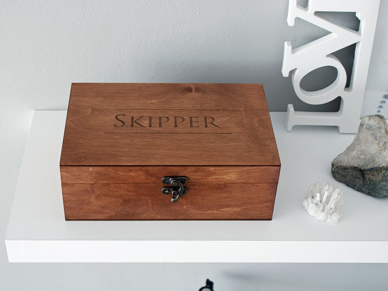 May include: A wooden box with a hinged lid and a metal clasp. The box is stained a dark brown colour and has the word "SKIPPER" engraved on the top.