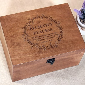 Op de afbeelding: Een houten doos met een scharnierend deksel en een metalen sluiting. De doos is gegraveerd met een krans van bladeren en de tekst "ELI SCOTT PLACKIS FOR THIS CHILD I PRAYED THE LORD HAS GRANTED THE DESIRES OF MY HEART" 1 SAMUEL 1:27.