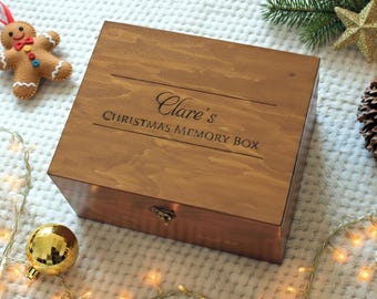 May include: A wooden memory box, inscribed with "Clare's Christmas Memory Box". The box is a warm brown colour and rectangular. A felt gingerbread man, gold bauble, and pine cone are placed nearby. The box has a metal clasp.