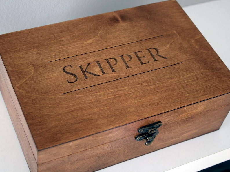 May include: A wooden box with a hinged lid and a metal clasp. The box is stained a dark brown colour and has the word "SKIPPER" engraved on the top.