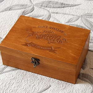 Puede incluir: Una caja de madera con tapa con bisagras y cierre de latón. La tapa está grabada con un pez y el texto "Alice Winslow - The Fishmaster".