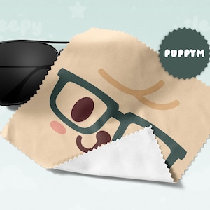May include: Black aviator sunglasses rest on a tan microfiber cloth with a cartoon face design. The cloth features teal-framed glasses, a brown nose, and a smiling mouth. A dark green circular logo with the text "PUPPYM" is also visible.