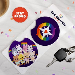Könnte beinhalten: Ein Set aus zwei runden Auto-Untersetzern auf einer weißen Trägerkarte mit der Aufschrift Car Coasters Set of 2. Ein Untersetzer zeigt einen Regenbogenstern mit Herz, der andere zeigt Zeichentricktiere mit Regenbogenflaggen.
