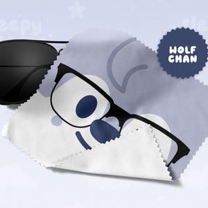 May include: Black aviator sunglasses rest on a white and blue microfiber cloth with a cartoon wolf face and the text "WOLF CHAN". The cloth is partially covering the sunglasses. The background is light blue with white stars.