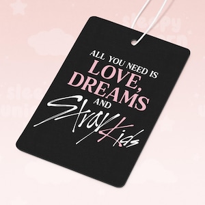 Puede incluir: Etiqueta rectangular negra con el texto "ALL YOU NEED IS LOVE, DREAMS AND Stray Kids" en letras blancas y rosas. La etiqueta tiene una cuerda blanca adjunta para colgar.