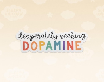 May include: A die-cut sticker featuring the black cursive text "desperately seeking" above the word "DOPAMINE" in bold, multicolored block letters including red, orange, yellow, green, blue, and purple against a white background.