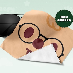 May include: Black aviator sunglasses rest on a tan cleaning cloth with a cartoon face design. The cloth features a pair of round glasses with maroon lenses and a red nose. A green circular logo with the text "HAN QUOKKA" is visible.