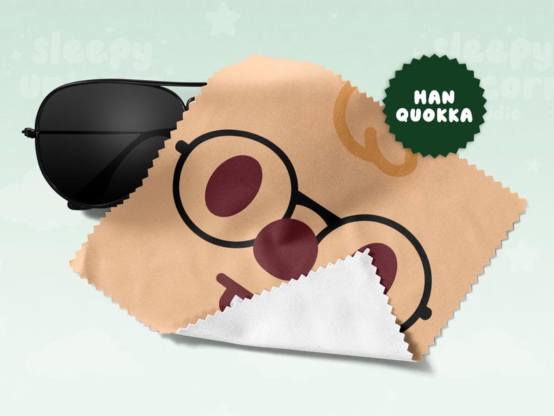May include: Black aviator sunglasses rest on a tan cleaning cloth with a cartoon face design. The cloth features a pair of round glasses with maroon lenses and a red nose. A green circular logo with the text "HAN QUOKKA" is visible.