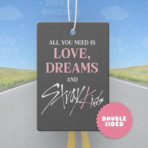 Könnte beinhalten: Ein rechteckiger, dunkelgrauer Lufterfrischer zum Aufhängen mit dem weißen Text ALL YOU NEED IS, dem rosa Text LOVE, DREAMS, dem weißen Text AND und dem weiß-rosa Text Stray Kids. Ein rosa, gezacktes, kreisförmiges Abzeichen zeigt DOUBLE SIDED.