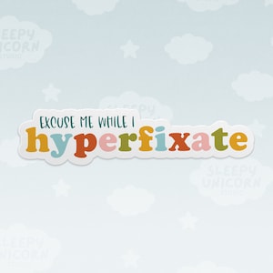 May include: A white sticker with the text "EXCUSE ME WHILE I hyperfixate" in colourful letters. The letters are in different colours, including orange, red, pink, blue, and green.