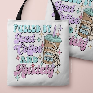 May include: White tote bag with black handles featuring the text "Fueled by Iced Coffee and Anxiety" in pastel colors. The design includes a cartoon iced coffee cup held by a skeleton hand, with star accents. The bag is perfect for carrying books, groceries, or everyday essentials.