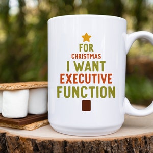 May include: White ceramic mug with the text "FOR CHRISTMAS I WANT EXECUTIVE FUNCTION" in green, orange, and brown. A star and square graphic are included in the design. A s'more is next to the mug.