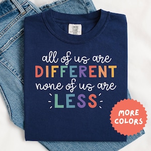 Könnte beinhalten: Marineblaues T-Shirt mit dem Text "all of us are DIFFERENT none of us are LESS" in bunten Buchstaben. Das Shirt wird auf einer blauen Jeans präsentiert. Ein roter Kreis mit den Worten "MORE COLORS" befindet sich unten rechts.