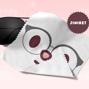 May include: Black aviator sunglasses rest next to a white cleaning cloth with a cartoon glasses design. A maroon circular sticker with the text "JINIRET" is also visible. The background is a soft pink.