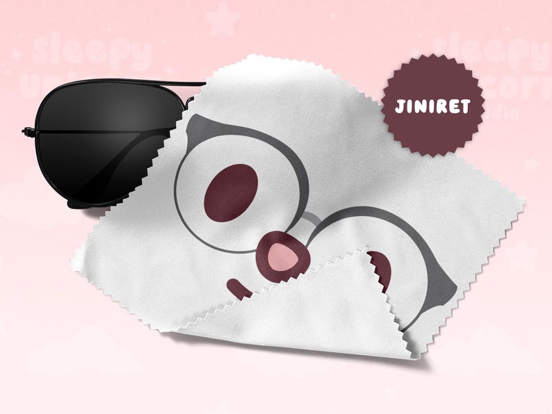 May include: Black aviator sunglasses rest next to a white cleaning cloth with a cartoon glasses design. A maroon circular sticker with the text "JINIRET" is also visible. The background is a soft pink.