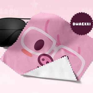 May include: Black aviator sunglasses rest on a pink microfiber cleaning cloth. The cloth features a cartoon pig wearing glasses. A dark purple circular badge with the text "DWAEKKI" is on the cloth. The background is a soft pink.