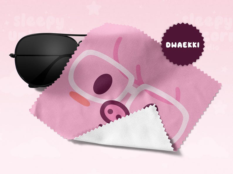May include: Black aviator sunglasses rest on a pink microfiber cleaning cloth. The cloth features a cartoon pig wearing glasses. A dark purple circular badge with the text "DWAEKKI" is on the cloth. The background is a soft pink.