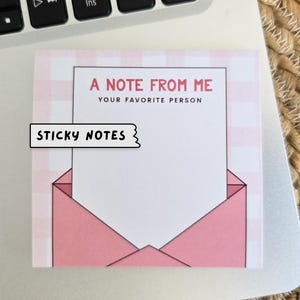 Könnte beinhalten: Ein Haftnotizblock mit rosa und weißem Karomuster, einem rosa Briefumschlagdesign und dem Text "A Note From Me Your Favorite Person" und "Sticky Notes" in Schwarz.