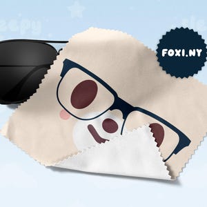 May include: Black aviator sunglasses next to a beige microfiber cleaning cloth with a cartoon glasses design. A navy blue circular logo with the text "FOXI.NY" is also visible.