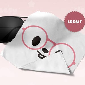May include: Black aviator sunglasses rest on a white cleaning cloth with a pink glasses graphic. A maroon circular sticker with the text "LEEBIT" is in the upper right corner. The background is a soft pink.