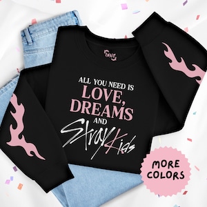 Könnte beinhalten: Schwarzes Sweatshirt mit dem Text "ALL YOU NEED IS LOVE, DREAMS AND Stray Kids" in Rosa und Weiß. Auf den Ärmeln befinden sich rosa Flammenmuster. Das Sweatshirt ist über einer Jeans gefaltet.