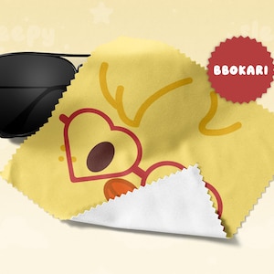 May include: Black aviator sunglasses rest on a yellow cleaning cloth with a heart-shaped glasses design. The cloth has a white edge and a red circular sticker with the text "BBOKARI".