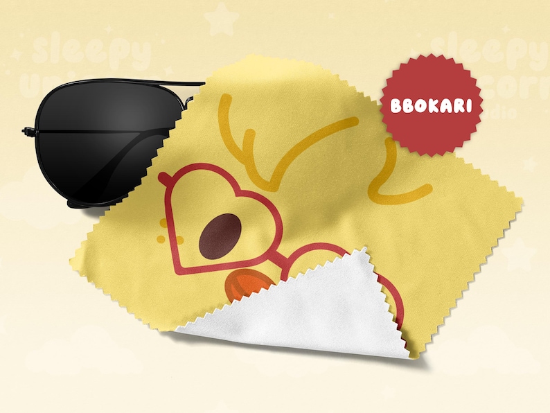 May include: Black aviator sunglasses rest on a yellow cleaning cloth with a heart-shaped glasses design. The cloth has a white edge and a red circular sticker with the text "BBOKARI".