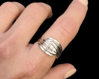 Puede incluir: Un anillo plateado con un diseño en capas en forma de ola. El anillo se lleva en un dedo sobre un fondo negro. La superficie del anillo refleja la luz, resaltando sus detalles texturizados. La joya es un accesorio simple pero elegante.