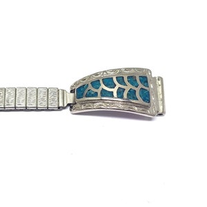 Peut inclure: Un bracelet de montre argenté avec un fermoir rectangulaire orné. Le fermoir présente une incrustation turquoise avec un motif en forme de feuille. Le bracelet est fait de petits maillons rectangulaires et connectés.