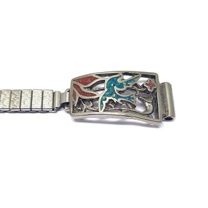 May include: A silver-toned watch band with an ornate rectangular clasp. The clasp features a bird design with red and turquoise accents. The band is made of small, connected metal links.