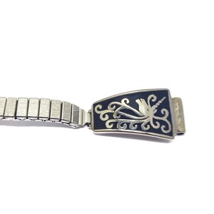 May include: A silver watch band with a black and silver clasp. The clasp features a decorative design of a stylized bird with a floral pattern.