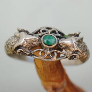 Könnte beinhalten: Ein silberner Ring mit zwei Wolfsköpfen, die sich gegenüberstehen, und einem smaragdgrünen Edelstein in der Mitte. Der Ring hat aufwendige Details und wird auf einem Holzstab präsentiert.