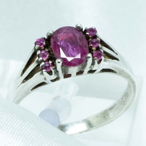 Peut inclure: Bague en argent ornée d'un rubis rose foncé ovale au centre, entouré de petits rubis assortis. La bague présente un design à tige fendue. Le bijou est présenté sur un fond de satin blanc.