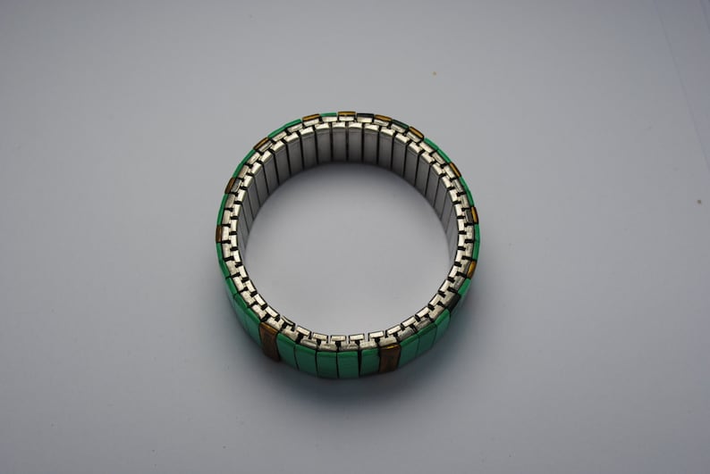 May include: A green and gold expandable bracelet made of metal links. The bracelet is made of small, rectangular pieces of metal that are connected together to form a flexible band.
