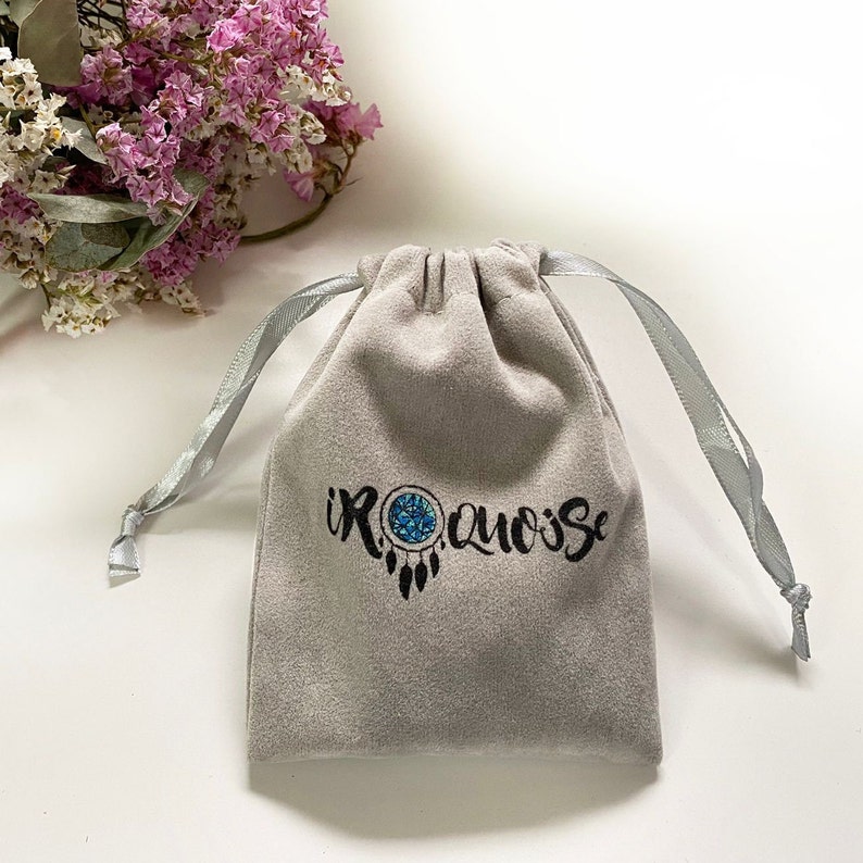 May include: A light gray velvet drawstring bag with a dreamcatcher design and the text "iROQUOISE" in black. The dreamcatcher has a blue center. The bag has a silver ribbon drawstring and is on a white surface.