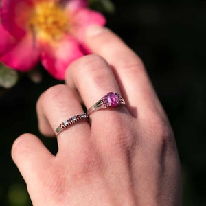 May include: Two silver rings with gemstones. One ring features a large oval pink gemstone surrounded by smaller pink stones. The other ring has a row of small red and white gemstones. The rings are on a hand, with a pink flower in the background.