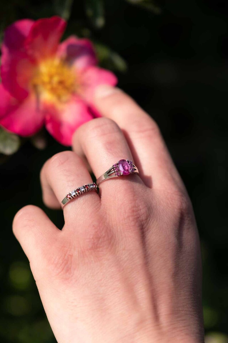 May include: Two silver rings with gemstones. One ring features a large oval pink gemstone surrounded by smaller pink stones. The other ring has a row of small red and white gemstones. The rings are on a hand, with a pink flower in the background.