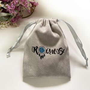May include: A gray velvet pouch with a drawstring closure and a silver ribbon. The pouch has a black embroidered logo that says "IROQUOISE" with a dreamcatcher design.
