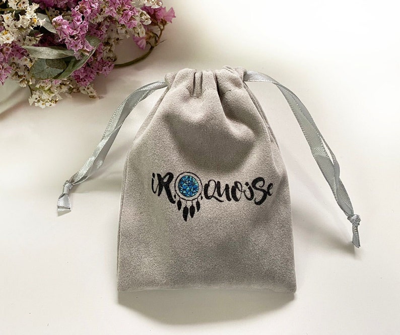 May include: A gray velvet pouch with a drawstring closure and a silver ribbon. The pouch has a black embroidered logo that says "IROQUOISE" with a dreamcatcher design.