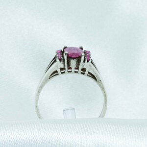 May include: A silver ring featuring a large oval pink gemstone, flanked by smaller pink stones. The ring has a detailed band with a split design. The setting and band appear to be made of a light-colored metal.