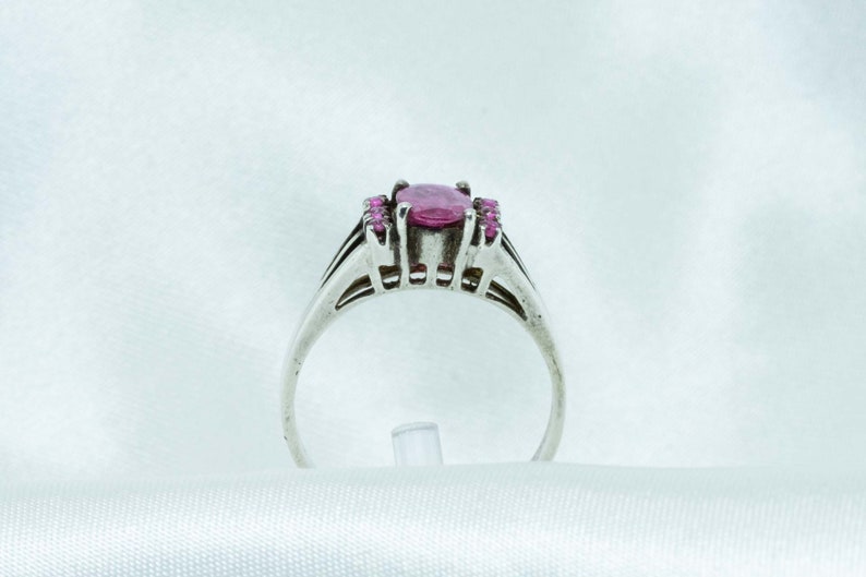 May include: A silver ring featuring a large oval pink gemstone, flanked by smaller pink stones. The ring has a detailed band with a split design. The setting and band appear to be made of a light-colored metal.