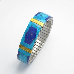 May include: A stretch bracelet featuring alternating segments of bright turquoise and deep blue stone inlays. Two gold-toned metal rectangular accents are integrated into the design, one engraved with an arrow and the other with a feather motif.